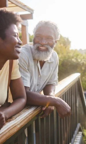 Couple laughing while standing on the balcony of their holiday rental and looking at the scenic view during a vacation